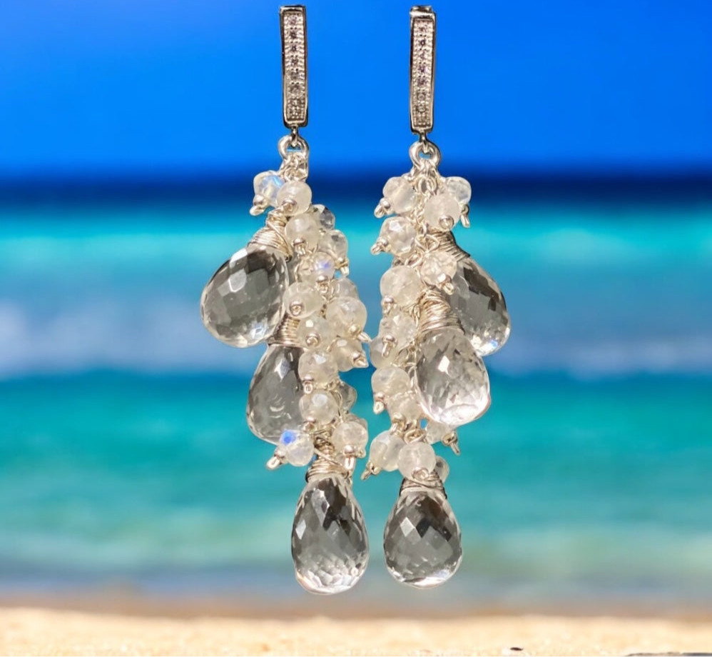Pair of dangling earrings with clear stones against a blurred beach background
