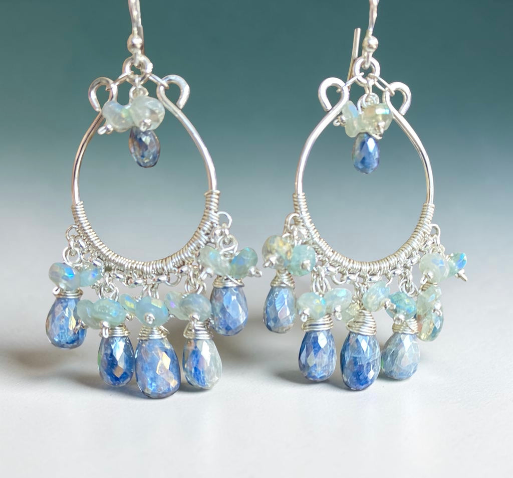 A pair of handmade silver hoop dangle earrings decorated with faceted blue and iridescent beads, shown against a soft blue gradient background.
