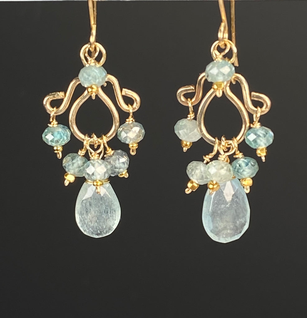 Gold-filled chandelier earrings hanging against a black background, highlighting pale blue moss aquamarine drops and sparkling blue zircon accents.

