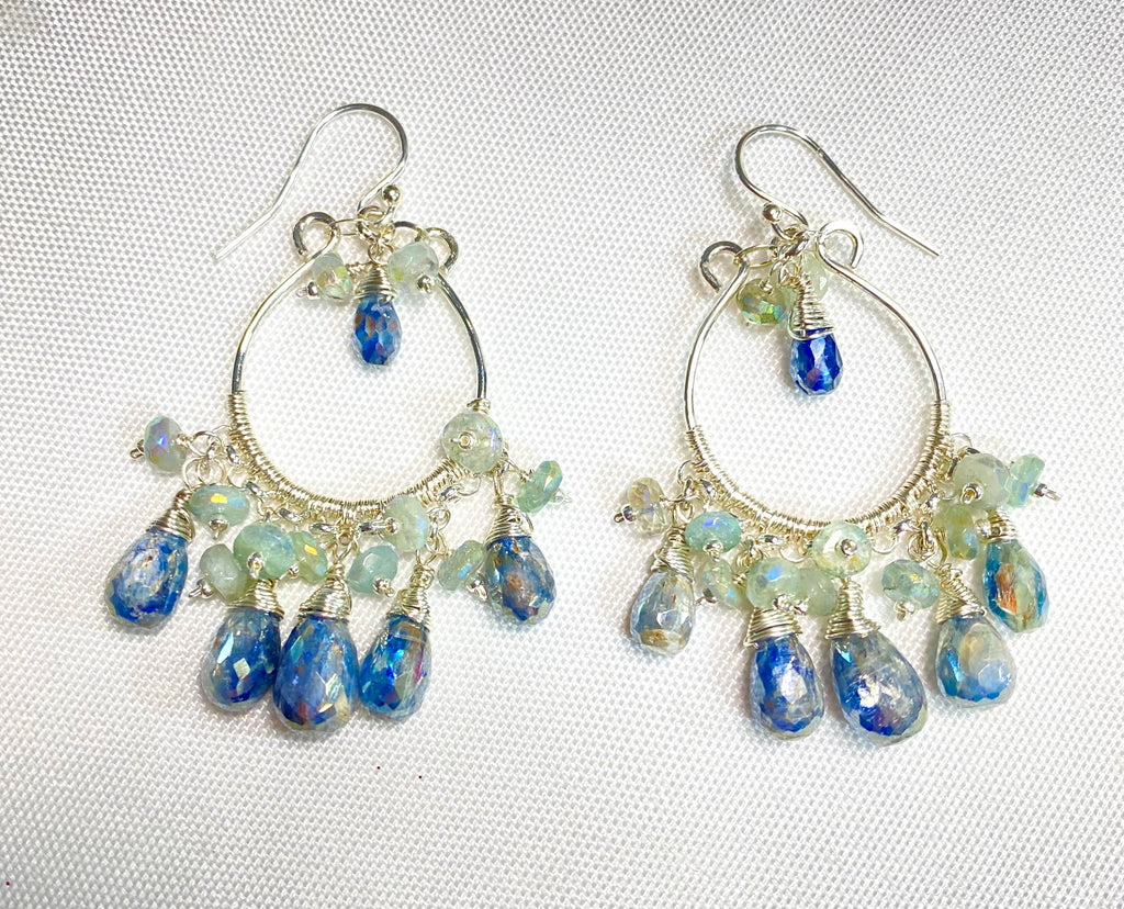 A pair of sterling silver hoop chandelier earrings with faceted blue teardrop beads and iridescent light blue beads, shown agains a textured white background.