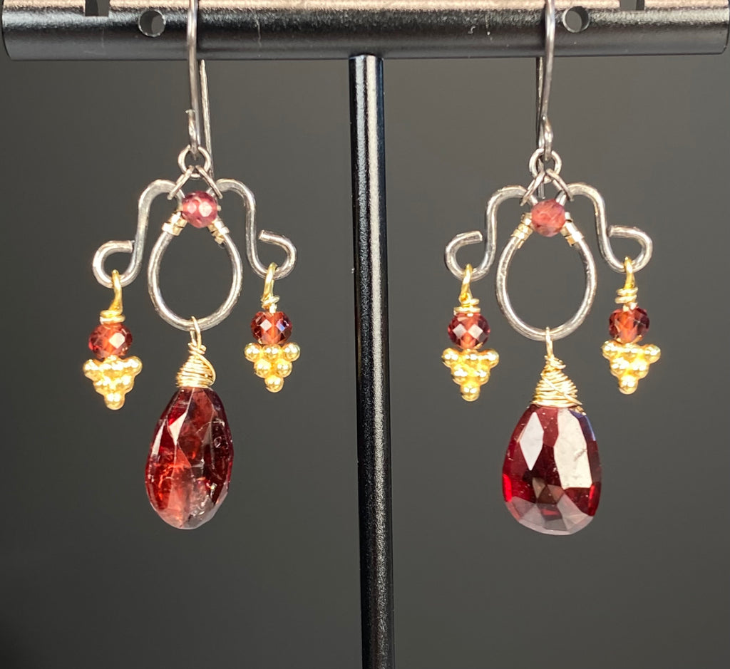 Red garnet chandelier earrings hanging from a metal display stand, showing French hooks, curled silver frames, and gold-wrapped pear drops.