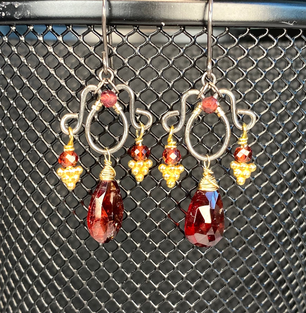 Hand-forged mixed-metal garnet chandelier earrings against metal mesh, showing faceted red stones and granulated gold vermeil details.