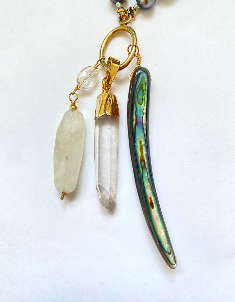 A close-up of a jewelry charm or pendant featuring three hanging elements: a pale milky white bead, a clear quartz crystal point with gold cap, and a curved iridescent abalone shell piece with gold hardware, all on a white background.