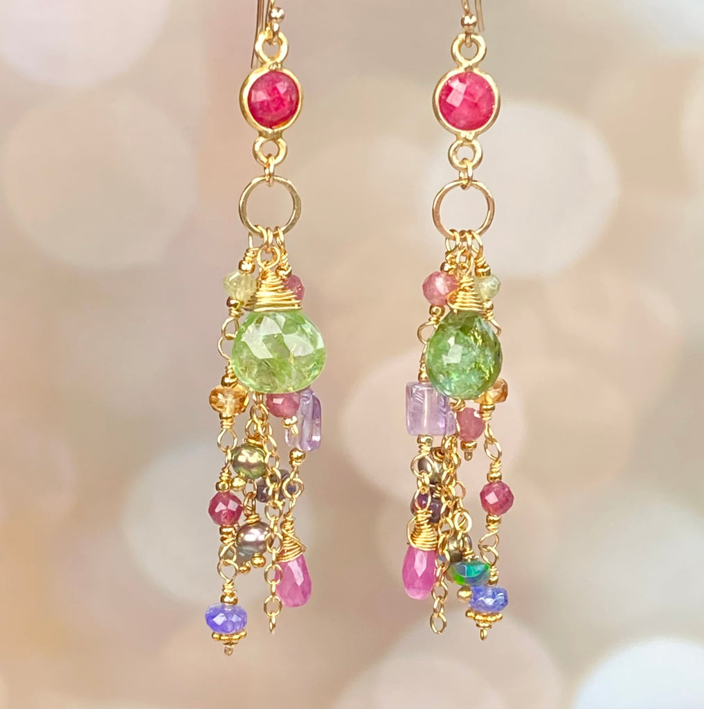 A pair of gold dangle earrings with faceted gemstones, featuring a round pink stone at the top, a larger green stone, and smaller dangling beads in pink, purple, and gold against a soft beige bokeh background.