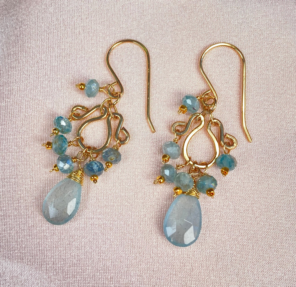 wider view on satin:
Pair of moss aquamarine and blue zircon earrings laid on pale satin, with gold-filled teardrop-and-scroll forms and dangling faceted blue stones.
