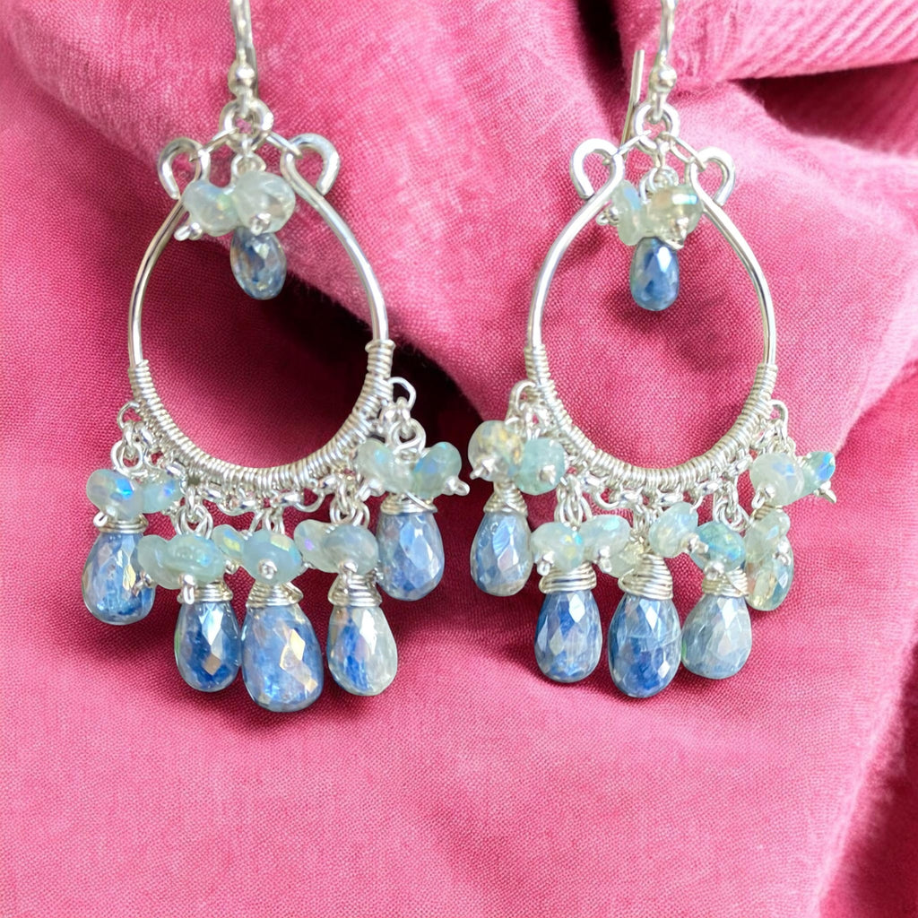 A pair of sterling silver hoop chandelier earrings with faceted light blue teardrop beads and iridescent blue-green stones, displayed on a textured pink fabric background.