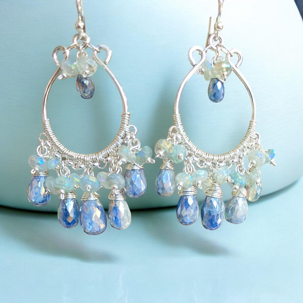 A pair of sterling silver hoop chandelier earrings with faceted blue teardrop beads and iridescent light blue beads, displayed against a light blue fabric background.