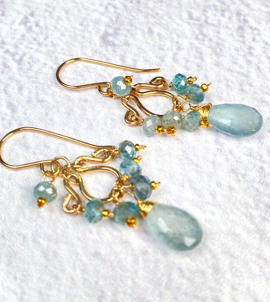 Angled view of moss aquamarine and blue zircon earrings on textured white fabric, showing gold-filled wire wrapping and faceted aquamarine drops.