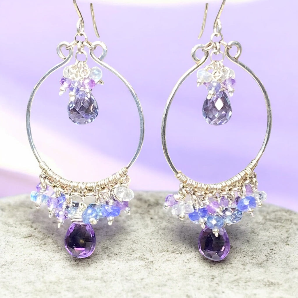 Colorful gemstone chandelier earrings amethyst tanzanite blue violet quartz dangling over grey mottled marble base with lavender backdrop
