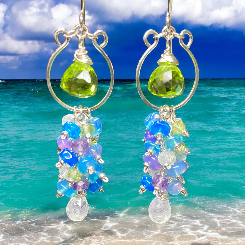 Close-up of teal, baby blue and lavender opal rondelles with peridot and moonstone on sterling silver chandelier earrings against beach surf background