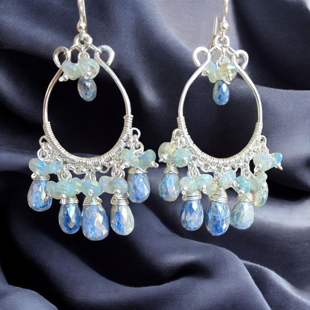 A pair of sterling silver hoop chandelier earrings with faceted light blue and iridescent teardrop beads, displayed on a dark navy blue satin fabric background.