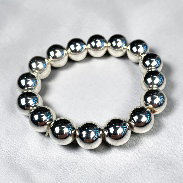 Silver hematite stretch bracelet with polished 12 mm round beads arranged in a circle on a light-grey fabric background.