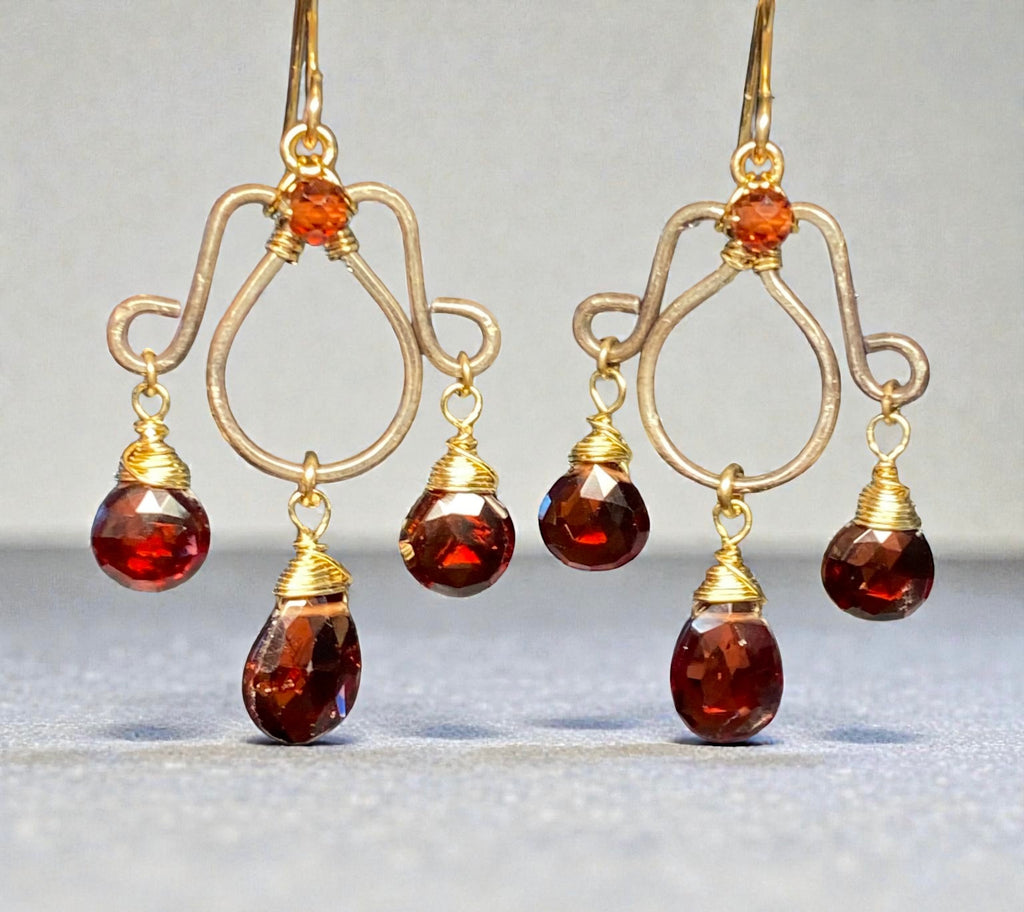 Oxidized silver chandelier earrings dangling red garnet faceted briolettes wire wrapped in 14kt gold fill wires shown in front of a two tone grey background.