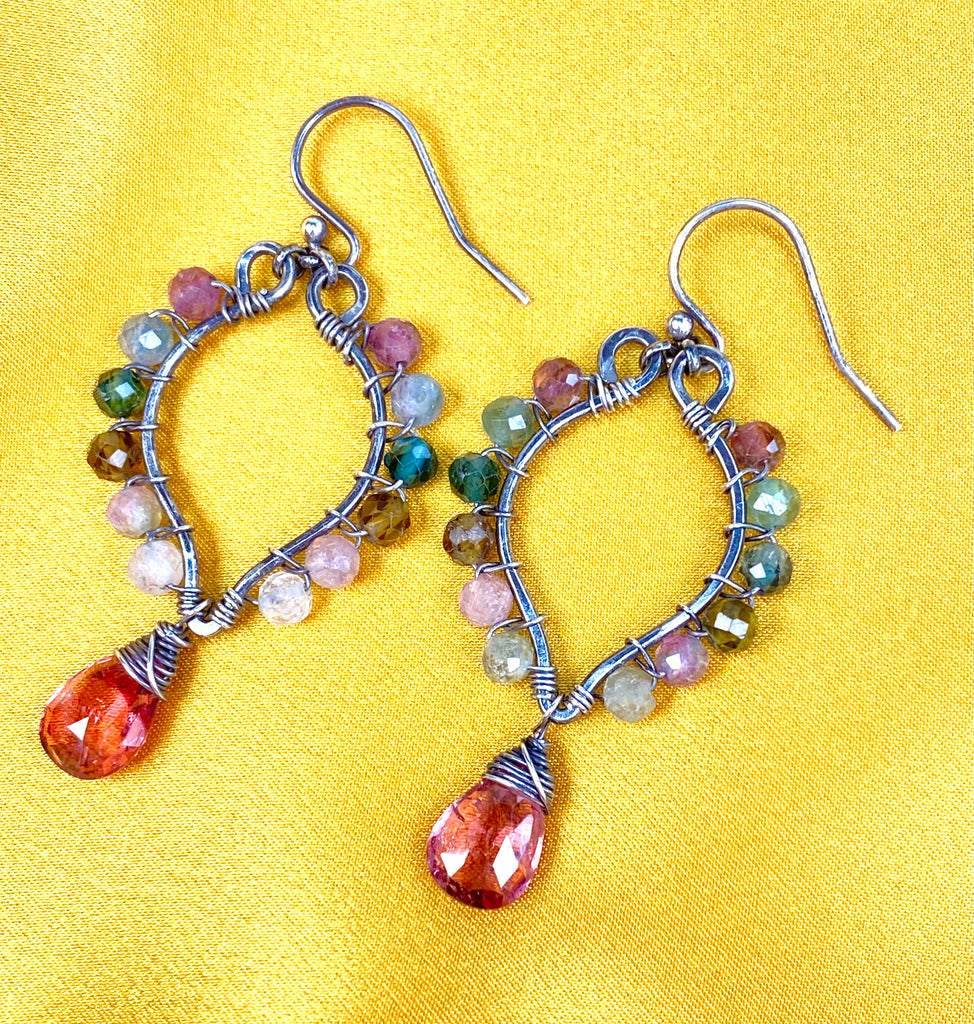 A pair of handmade wire-wrapped dangle hoop earrings with faceted multicolor tourmaline beads and largepink tourmaline teardrop gemstones, displayed on a bright yellow fabric background.