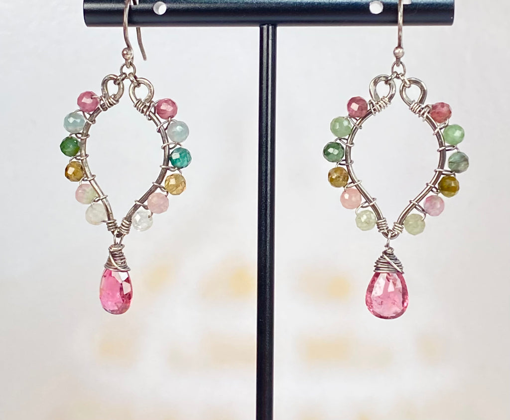 A pair of handmade hoop earrings displayed on a black stand. The earrings feature an oxidized silver-gunmetal tone wire frame holding small multicolored tourmaline gemstone beads in pink, green, yellow, blue and white, with a dangling pink tourmaline teardrop gemstone at the bottom.