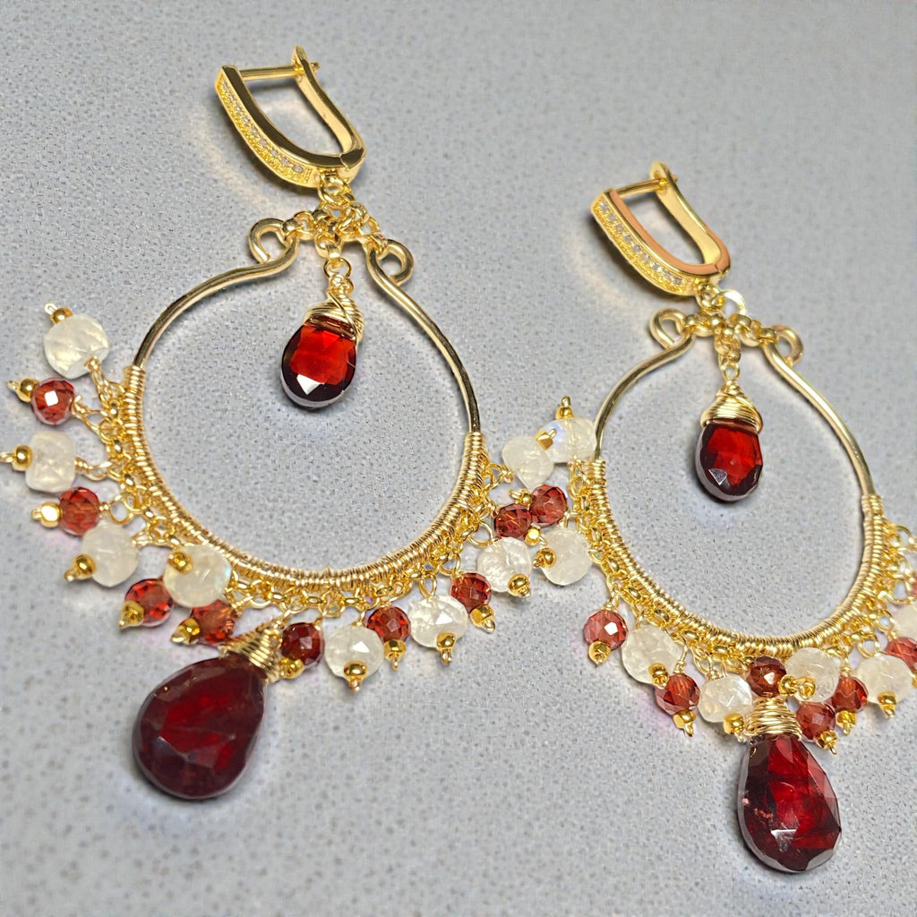 Red garnet gold hoop earrings with rainbow moonstone lying flat on light grey background