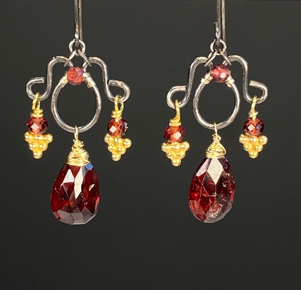 Red garnet chandelier earrings against a dark background, showing oxidized silver frames, faceted garnet pear drops, and golden granulated accents.