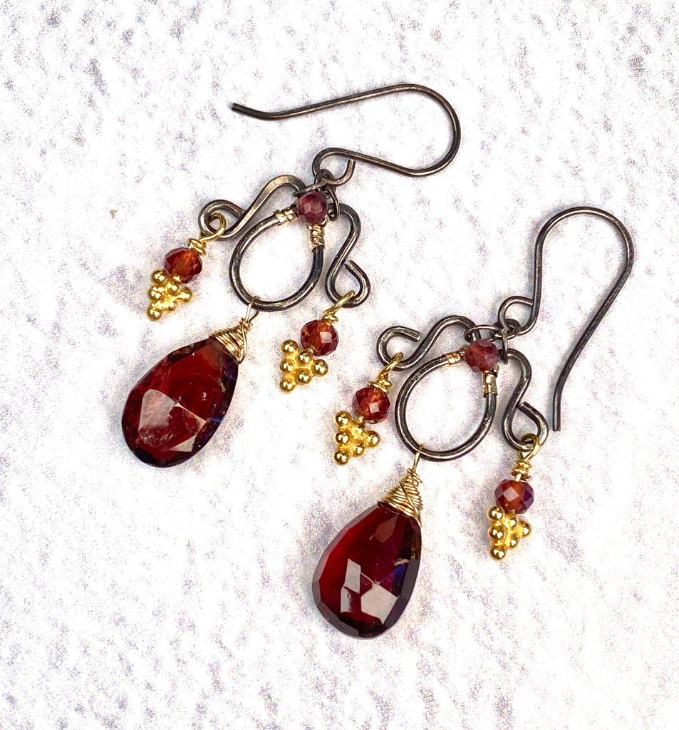 Oxidized sterling silver chandelier earrings resting on white textured fabric, with deep red garnet briolettes and gold accents.
