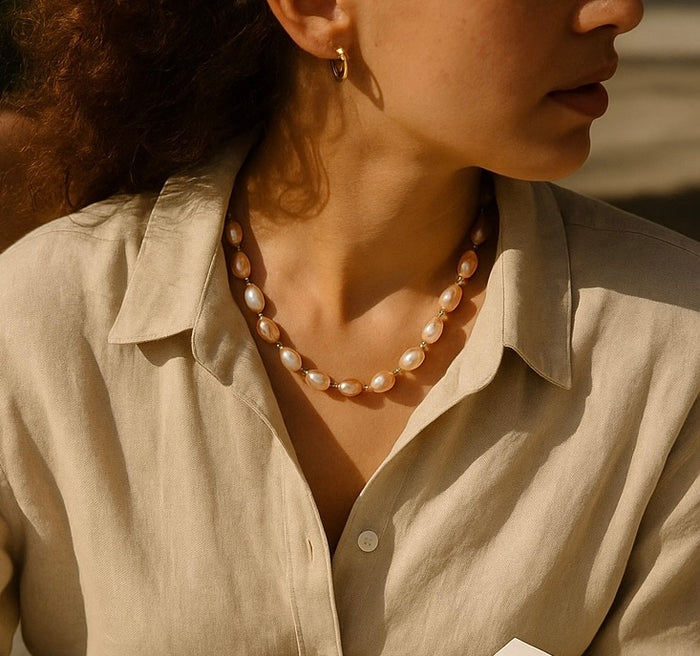 Styling image of a metallic baroque pearl and spinel necklace worn with an open-collar neutral blouse.
