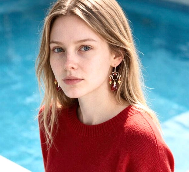 Model wearing red garnet chandelier earrings with oxidized silver frames and gold accents, paired with a red sweater beside a pool.