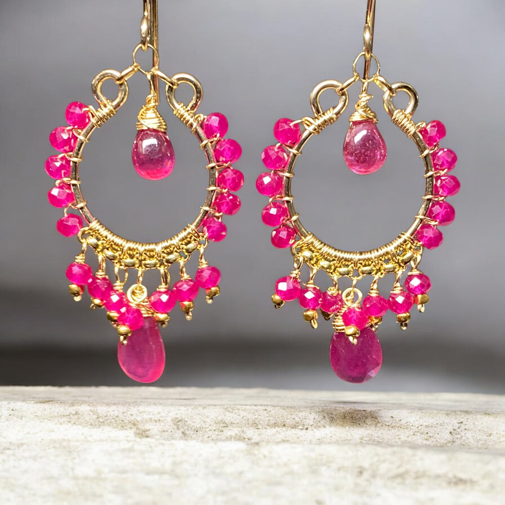 Red ruby pink sapphire gold chandelier hoop earrings dangling over mottled marble slab with grey background