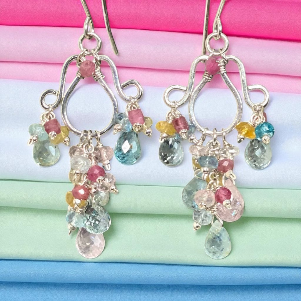Multicolor aquamarine pink tourmaline chandelier earrings with pastel blue pink yellow gemstones dangling in front of stack of folded pink white and mint green fabrics