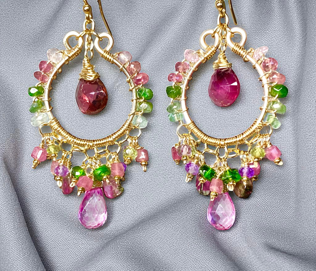 Gold hoop earrings with pink and green gemstones on a gray fabric background